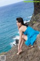 A woman in a blue dress sitting on a rock by the ocean.